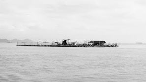 A solitary floating structure positioned in a vast body of water, possibly an aquaculture platform or fisherman's hut. The horizon features distant landmasses and ships, under a cloudy sky.