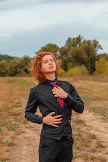 A person with curly red hair stands confidently in an outdoor setting, wearing a black suit with a maroon tie. The background is a natural landscape with trees and an open field.