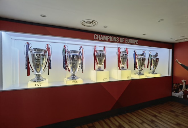 A display case featuring six silver trophies with ribbons, each marked with a year: 1977, 1978, 2005, 2019, 1981, and 1984. Above the case is a sign reading 'Champions of Europe'. The trophies are evenly spaced and lit from below, set against a red and white backdrop. A few people can be partially seen in the bottom right corner.