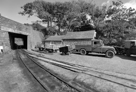 A black and white photograph features a railway track leading into a tunnel on the left side of the image. In the center, there is a small complex of structures including a building with a sloped roof and exposed brick walls. Multiple carts and two vintage trucks are stationed near the building. Lush trees and foliage are visible in the background, providing a natural setting for the scene.