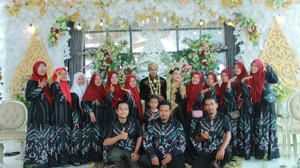 A group of people dressed in coordinated outfits are posing in a decorated space. The backdrop features abundant floral arrangements in various colors including white, green, and pink. The individuals in the back row are predominantly women wearing long dresses with matching patterns and red headscarves. In the center of the back row are a couple, possibly the bride and groom, wearing traditional wedding attire. Seated in the front row are men and a child, also dressed in matching patterned shirts.