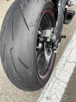A close-up view of a motorcycle rear tire showing significant wear and rubber debris build-up from usage. The tire features a tread pattern designed for road use and is accompanied by a visible disc brake system.