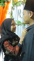 A woman in a black hijab is looking up and smiling at a man wearing a black hat. She is adjusting his collar with her hands. They are standing close to each other in front of a white and green floral backdrop with an 'R' in the center. An orange draped cloth is visible on the left.