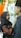 A woman in a black hijab is looking up and smiling at a man wearing a black hat. She is adjusting his collar with her hands. They are standing close to each other in front of a white and green floral backdrop with an 'R' in the center. An orange draped cloth is visible on the left.