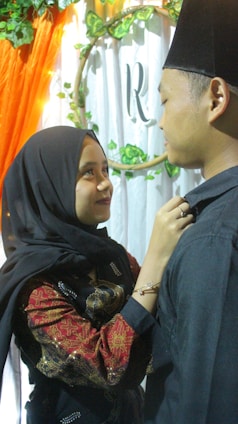 A woman in a black hijab is looking up and smiling at a man wearing a black hat. She is adjusting his collar with her hands. They are standing close to each other in front of a white and green floral backdrop with an 'R' in the center. An orange draped cloth is visible on the left.