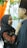 A woman in a black hijab is looking up and smiling at a man wearing a black hat. She is adjusting his collar with her hands. They are standing close to each other in front of a white and green floral backdrop with an 'R' in the center. An orange draped cloth is visible on the left.