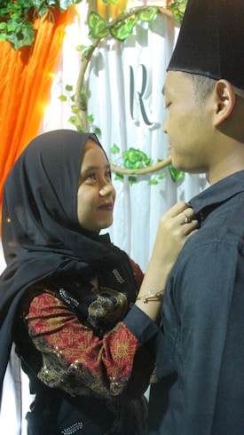 A woman in a black hijab is looking up and smiling at a man wearing a black hat. She is adjusting his collar with her hands. They are standing close to each other in front of a white and green floral backdrop with an 'R' in the center. An orange draped cloth is visible on the left.