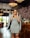 A stylish woman in a geometric patterned dress is standing inside a chic, modern restaurant or cafe. She is wearing sunglasses and is smiling confidently. The setting features elegant chandeliers, decorative lighting, and plush seating, with a floral decoration in the background.