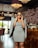 A stylish woman in a geometric patterned dress is standing inside a chic, modern restaurant or cafe. She is wearing sunglasses and is smiling confidently. The setting features elegant chandeliers, decorative lighting, and plush seating, with a floral decoration in the background.