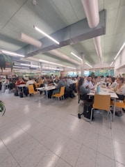 A spacious cafeteria filled with people sitting at tables. The ceiling features modern lighting and ventilation systems. The chairs are yellow, contrasting with the tiled floor and light green ceiling. Groups of people are engaged in conversation or working, indicating a lively atmosphere.