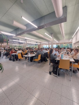 A spacious cafeteria filled with people sitting at tables. The ceiling features modern lighting and ventilation systems. The chairs are yellow, contrasting with the tiled floor and light green ceiling. Groups of people are engaged in conversation or working, indicating a lively atmosphere.