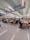 A spacious cafeteria filled with people sitting at tables. The ceiling features modern lighting and ventilation systems. The chairs are yellow, contrasting with the tiled floor and light green ceiling. Groups of people are engaged in conversation or working, indicating a lively atmosphere.