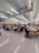 A spacious cafeteria filled with people sitting at tables. The ceiling features modern lighting and ventilation systems. The chairs are yellow, contrasting with the tiled floor and light green ceiling. Groups of people are engaged in conversation or working, indicating a lively atmosphere.