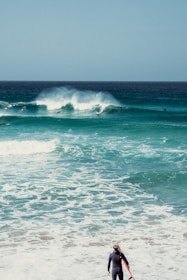A surfer in a wetsuit stands on the shore, holding a surfboard, while observing the ocean waves. Several surfers are visible riding the waves in the background under a clear sky.