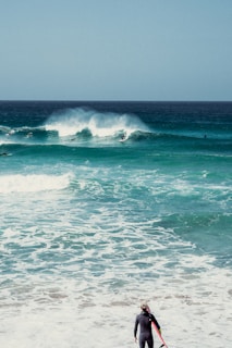 A surfer in a wetsuit stands on the shore, holding a surfboard, while observing the ocean waves. Several surfers are visible riding the waves in the background under a clear sky.