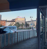 An urban street scene featuring several multi-story buildings in the background, with a prominent advertisement for 'Tolle Angebote'. Cars are visible on the road, and a sidewalk with lampposts runs alongside. The image is taken from a covered area, possibly a station or bus stop, providing a framing effect.