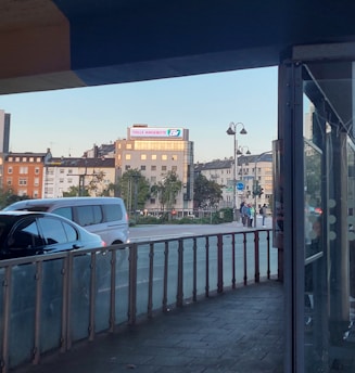 An urban street scene featuring several multi-story buildings in the background, with a prominent advertisement for 'Tolle Angebote'. Cars are visible on the road, and a sidewalk with lampposts runs alongside. The image is taken from a covered area, possibly a station or bus stop, providing a framing effect.