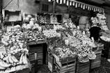 A fruit and vegetable market stall displays a variety of produce, including bananas, melons, grapes, and citrus fruits, all arranged in wooden crates. Price tags are visible on some of the boxes, indicating the cost of the items. A person with a cap is standing on the right side, presumably a vendor or customer.