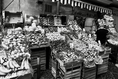 A fruit and vegetable market stall displays a variety of produce, including bananas, melons, grapes, and citrus fruits, all arranged in wooden crates. Price tags are visible on some of the boxes, indicating the cost of the items. A person with a cap is standing on the right side, presumably a vendor or customer.