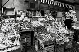 A fruit and vegetable market stall displays a variety of produce, including bananas, melons, grapes, and citrus fruits, all arranged in wooden crates. Price tags are visible on some of the boxes, indicating the cost of the items. A person with a cap is standing on the right side, presumably a vendor or customer.