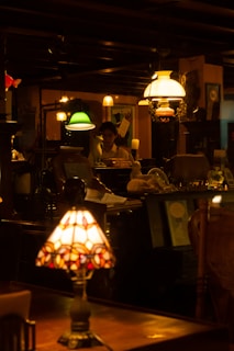A dimly lit interior of a vintage or antique store with several lit lamps in varied styles, including a stained-glass table lamp in the foreground. There are various objects, posters, and decorative elements throughout the room, creating a cozy and nostalgic atmosphere. A person is visible at the back, partially obscured by the items in the room.