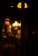 A dimly lit interior with warm amber lighting, featuring a variety of decorative items. A prominent statue of Buddha is visible in the background, surrounded by vintage signs, including an 'OPEN' sign and another sign with Chinese characters. An ornate chandelier and a large lamp contribute to the soft, inviting glow.