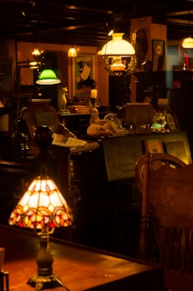 A dimly lit antique shop with various vintage objects, including lamps with stained glass and colored shades. Shelves are filled with decorative items and furniture, creating a nostalgic and cozy atmosphere.