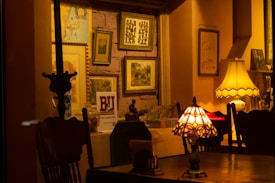 A warmly lit interior space featuring a collection of framed pictures on a wall, table lamps with intricate designs, and wooden furniture. The space has a cozy and vintage aesthetic with warm yellow and brown tones.