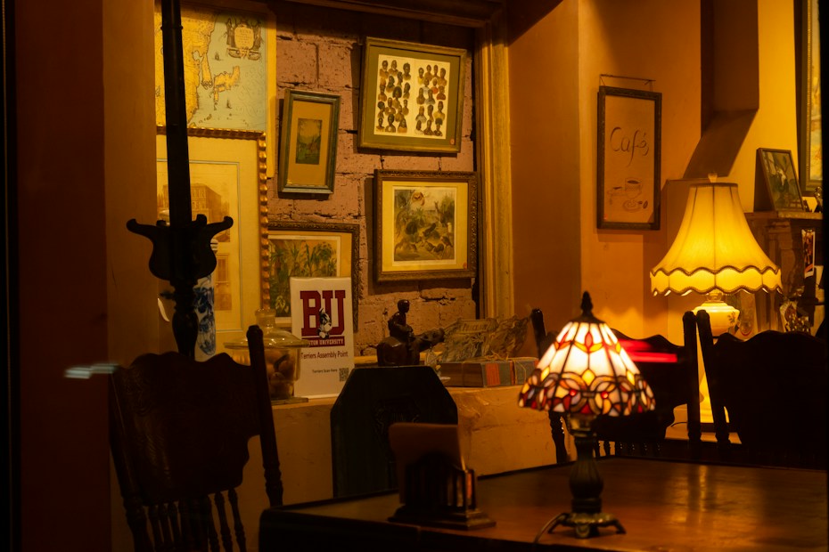 A warmly lit interior space featuring a collection of framed pictures on a wall, table lamps with intricate designs, and wooden furniture. The space has a cozy and vintage aesthetic with warm yellow and brown tones.