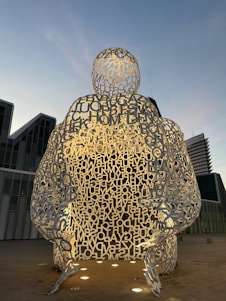 The image features a large sculpture made from metallic letters forming a hollow human figure, set outdoors against a backdrop of modern architecture. The sculpture is illuminated from below, creating a dramatic contrast with the early evening sky.
