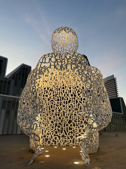 The image features a large sculpture made from metallic letters forming a hollow human figure, set outdoors against a backdrop of modern architecture. The sculpture is illuminated from below, creating a dramatic contrast with the early evening sky.