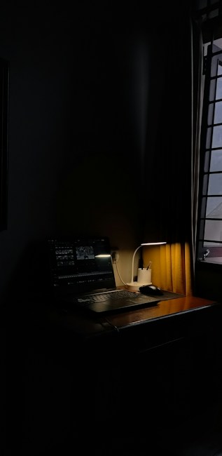 A dimly lit room features a wooden desk with a laptop and a desk lamp. Soft light casts shadows around the workspace, providing an intimate ambiance. Curtains partially cover a window, adding to the moody lighting.