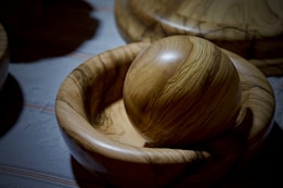 A smooth, spherical wooden object is resting inside a shallow wooden bowl. The wood has distinct, dark grain patterns running through it, highlighting a polished surface that catches the light. The background is a textured surface with muted colors, emphasizing the natural tones of the wood.