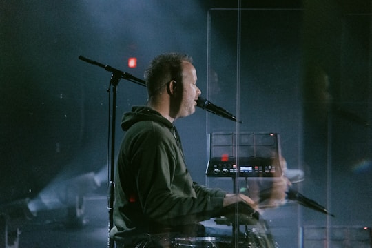 A person in a hoodie is seated at a keyboard or synthesizer with a microphone in front of them. The atmosphere is dimly lit, likely on a stage with some visible lighting equipment in the background. There is a clear reflection visible, suggesting a transparent divider or screen between the person and the camera.