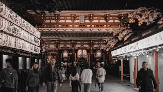 A group of people walk along a pathway leading to a traditional Japanese temple, illuminated with soft lighting. The temple features intricate wooden architecture and is adorned with red accents. On either side of the path are rows of lanterns with Japanese characters, and autumn leaves add a seasonal touch.