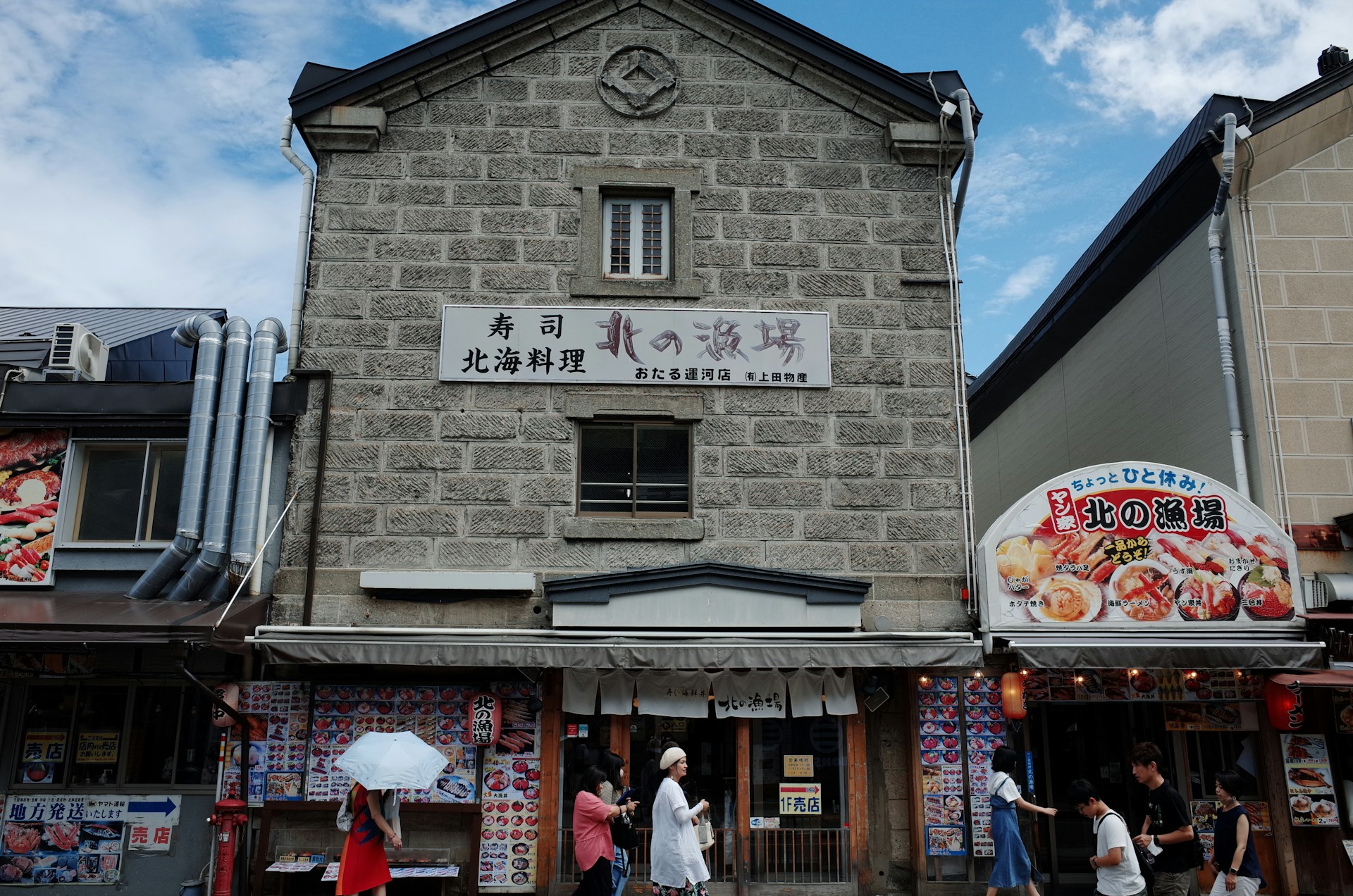 A stone building with Japanese signage features prominently, flanked by neighboring shops. There is visible text on the signs, promoting seafood and sushi. People are walking in front of the building, some browsing menus displayed outside, and one person holds a white umbrella. The sky is partly cloudy, providing a bright backdrop.