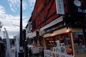 A bustling street market with shops and stalls displaying a variety of goods and signage in Japanese. The architecture is traditional with vibrant colors. People walk through the market area under a sky with scattered clouds.
