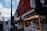 A bustling street market with shops and stalls displaying a variety of goods and signage in Japanese. The architecture is traditional with vibrant colors. People walk through the market area under a sky with scattered clouds.