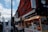 A bustling street market with shops and stalls displaying a variety of goods and signage in Japanese. The architecture is traditional with vibrant colors. People walk through the market area under a sky with scattered clouds.