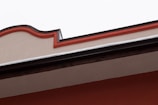 A section of a building's roof with a decorative edge featuring a wavy design. The top part of the wall is beige with a prominent reddish-orange border. Below the decorative edge, there is a layer of snow on the roof, and the lower part of the wall displays a muted red color.