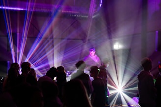 A DJ stands behind a console surrounded by colorful laser lights and silhouetted figures dancing. The atmosphere is vibrant with beams of purple, blue, and white light piercing through a smokey, dimly lit room, creating a lively and energetic environment.