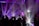 A DJ stands behind a console surrounded by colorful laser lights and silhouetted figures dancing. The atmosphere is vibrant with beams of purple, blue, and white light piercing through a smokey, dimly lit room, creating a lively and energetic environment.