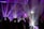 A DJ stands behind a console surrounded by colorful laser lights and silhouetted figures dancing. The atmosphere is vibrant with beams of purple, blue, and white light piercing through a smokey, dimly lit room, creating a lively and energetic environment.