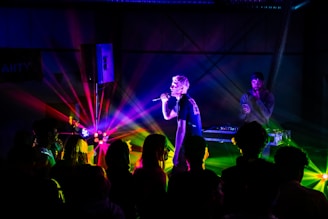 A lively concert scene with vivid, colorful lighting illuminating a performer holding a microphone. A DJ stands next to equipment in the background, while a crowd of people face the stage.