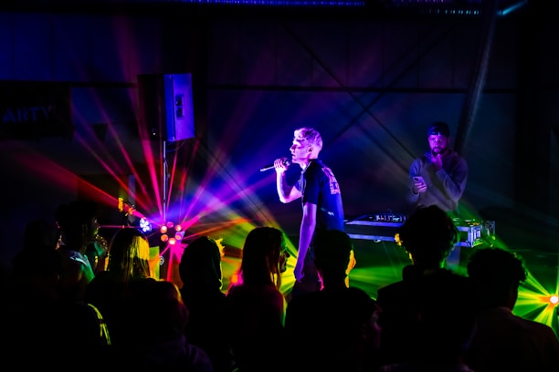 A lively concert scene with vivid, colorful lighting illuminating a performer holding a microphone. A DJ stands next to equipment in the background, while a crowd of people face the stage.