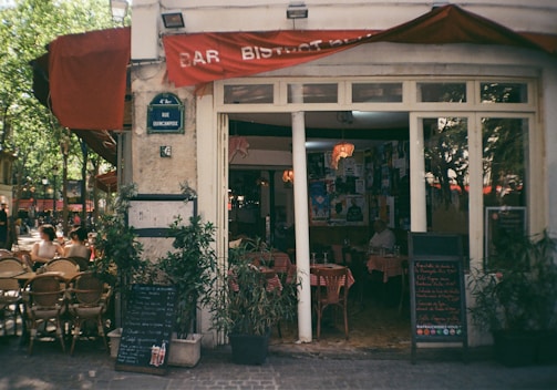 A quaint European bar bistro with red signage and a street sign indicating Rue Quincampoix. Wicker chairs and tables are set outside under a large red awning. Several people are sitting outside, enjoying the sunny weather. The interior of the bistro is visible through large windows, showing tables covered with red checkered tablecloths and walls adorned with various posters. Green potted plants flank the entrance, along with chalkboards displaying menus.