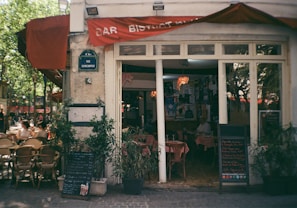 A quaint European bar bistro with red signage and a street sign indicating Rue Quincampoix. Wicker chairs and tables are set outside under a large red awning. Several people are sitting outside, enjoying the sunny weather. The interior of the bistro is visible through large windows, showing tables covered with red checkered tablecloths and walls adorned with various posters. Green potted plants flank the entrance, along with chalkboards displaying menus.