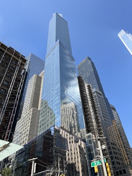 A group of modern skyscrapers with reflective glass facades, capturing the buildings surrounding them. The sky is clear, enhancing the brightness and clarity of the architectural details. The intersection of a city street is visible at the bottom, marked by a street sign and traffic lights.