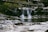 A tranquil scene of a small waterfall flowing into a shallow pool, surrounded by rocks and fallen branches. The setting appears to be in a forested area, with greenery and natural elements creating a serene atmosphere. The water cascades gently over moss-covered stones, creating a soothing natural sound.