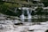 A tranquil scene of a small waterfall flowing into a shallow pool, surrounded by rocks and fallen branches. The setting appears to be in a forested area, with greenery and natural elements creating a serene atmosphere. The water cascades gently over moss-covered stones, creating a soothing natural sound.
